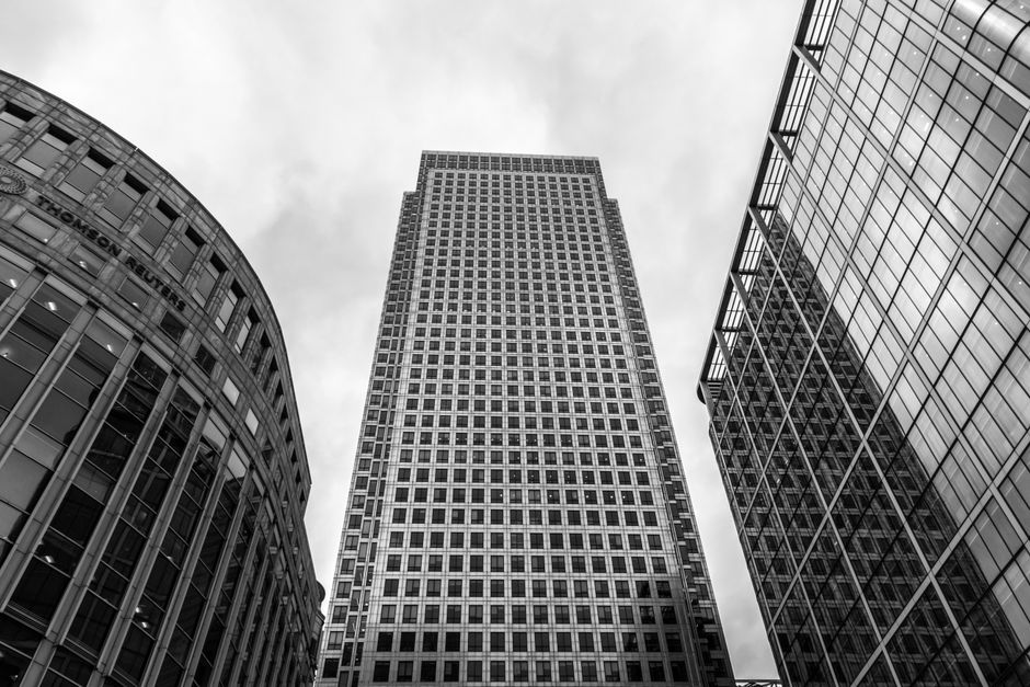 A black and white photograph taken from a low angle, capturing three modern high-rise office buildings in Canary Wharf. The building on the left features a curved façade with large glass windows and