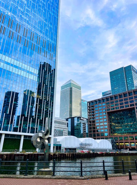 A wide view of the Heron Quays waterfront in Canary Wharf during daytime, featuring modern high-rise office buildings with glass and steel facades on both sides of the water. The calm water reflects t