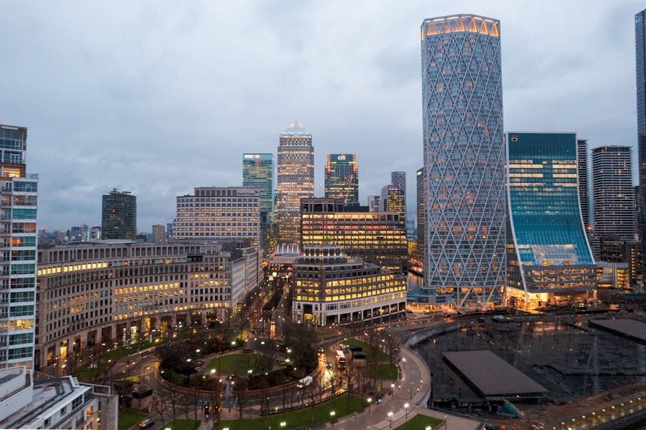A panoramic view of a modern city skyline with tall glass skyscrapers under a bright blue sky, visible through lush green trees and open parkland in the foreground, showcasing a combination of urban a