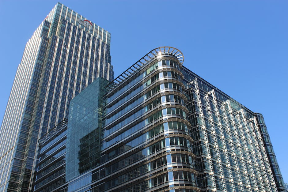 Interior view of a modern commercial building entrance with a large glass facade showing a cityscape of tall office buildings in Canary Wharf. The image features a revolving glass door with metal fram