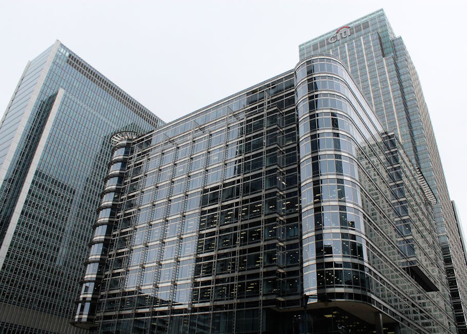 An aerial view of West India Quay in Canary Wharf showing a dense cluster of modern high-rise office buildings and residential towers with glass facades reflecting the cloudy sky. At the foreground, a